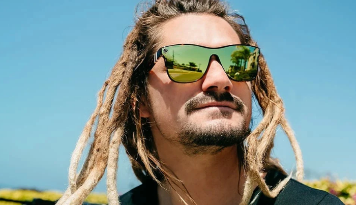 Person with dreadlocks wearing green sunglasses against a clear blue sky