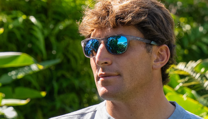 Man wearing Maui Jim sunglasses with a blue reflective surface, standing against a green leafy background