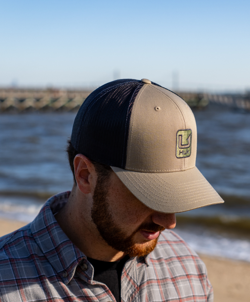 A man wearing a beige and navy trucker hat with a green and yellow logo patch on the front, photographed outdoors at the beach.