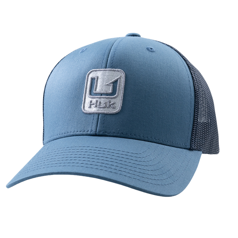 A blue trucker-style baseball cap with a structured front panel, curved brim, and dark mesh back panels. A square embroidered patch on the front displays a stylized 'H' logo above the word 'Huk', indicating the brand.
