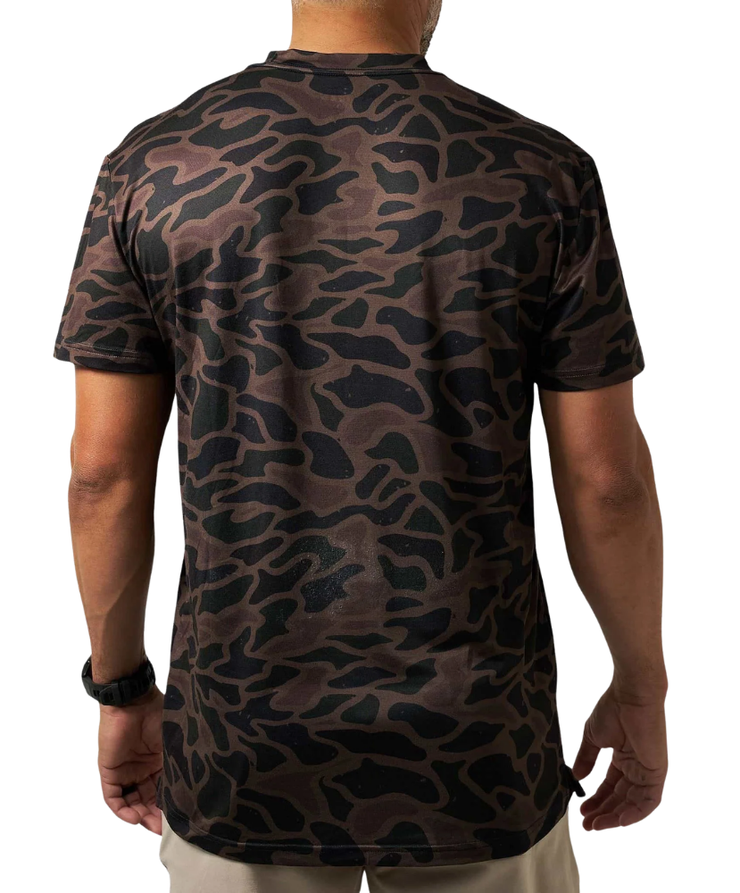 A man viewed from the back wearing a short-sleeve crewneck t-shirt with a dark brown and black organic camouflage pattern, paired with beige pants against a white background.