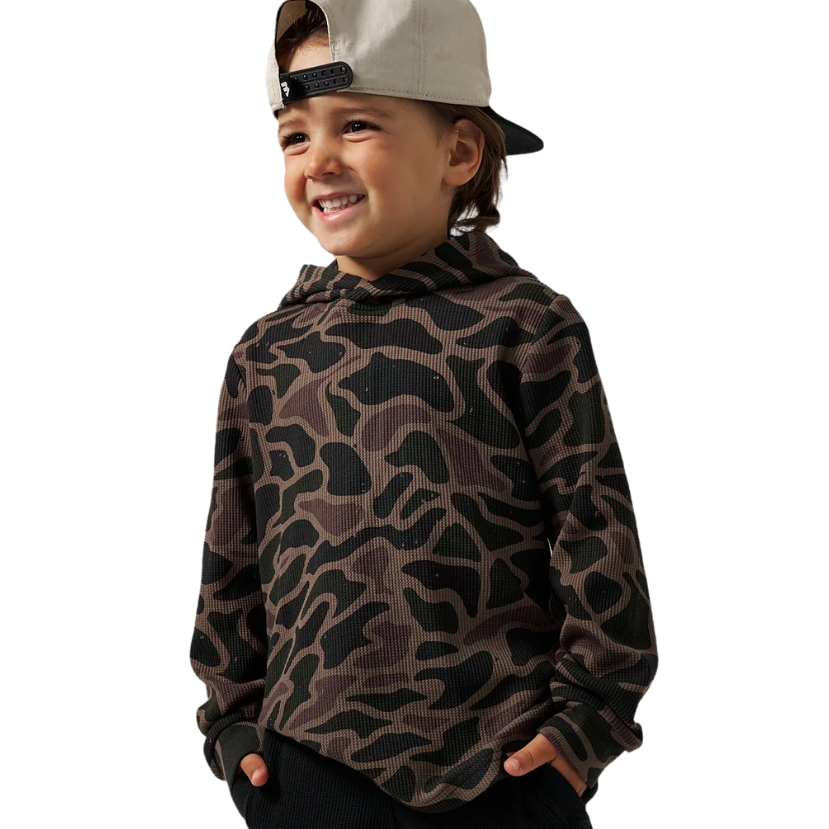A young boy wearing a brown and black camouflage patterned hoodie, black pants, and a light beige baseball cap, shown against a plain white background.