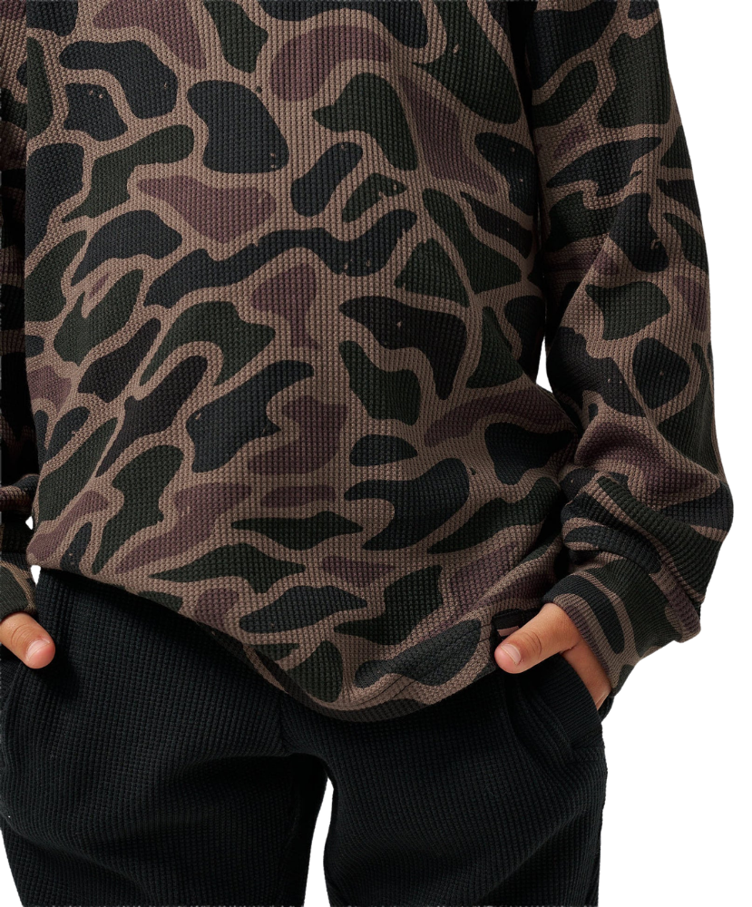 A close-up of a person wearing a long-sleeve waffle-knit top with a dark camouflage pattern in black, olive green, and brown, paired with black textured pants.