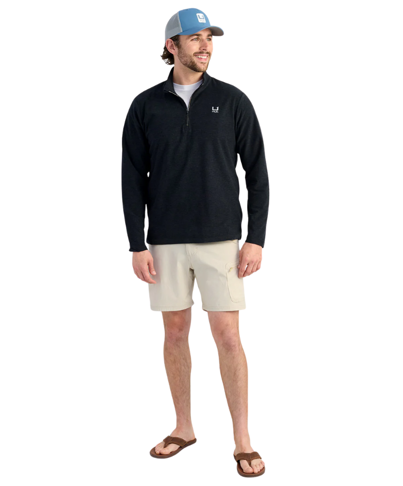 Man wearing a black long-sleeve HUK PULLOVER with beige shorts, and a blue cap on a white background