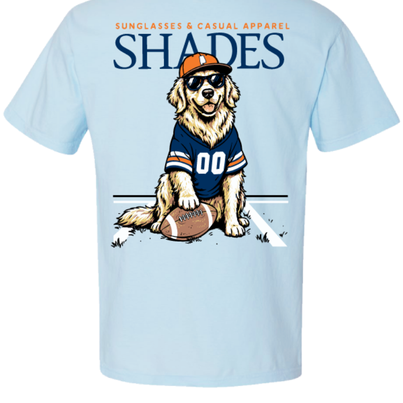 A light blue short-sleeve t-shirt shown from the back, featuring a graphic of a golden retriever wearing a navy and orange football jersey, sunglasses, and an orange cap, sitting next to a football. The word 'SHADES' is printed in large navy letters above the dog, with smaller orange text reading 'SUNGLASSES & CASUAL APPAREL' above that.