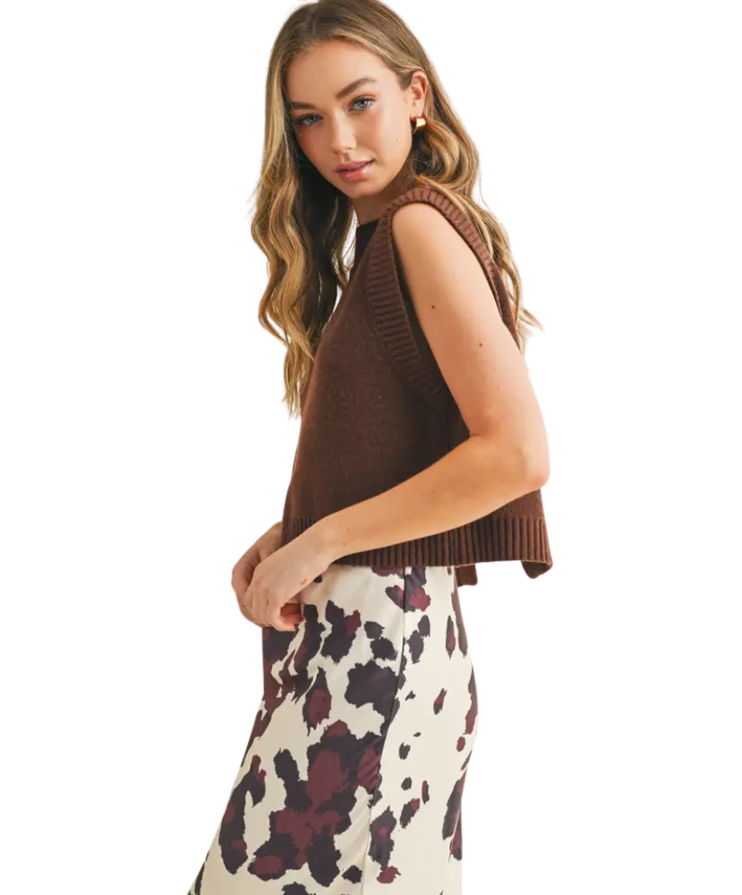 Woman wearing a brown sleeveless sweater over a floral dress on a white background