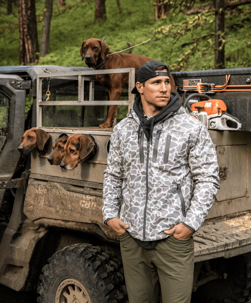 A man wearing a gray and white animal-print hooded jacket stands in front of a muddy off-road truck with dogs and a chainsaw in a forest setting.