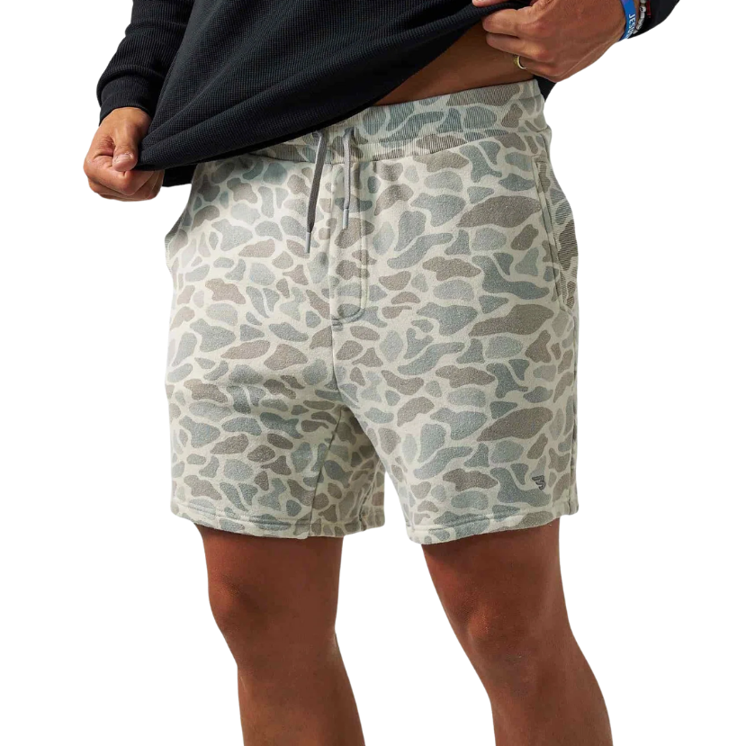 A person wearing patterned shorts and pulling up a black long-sleeve shirt against a plain white background.