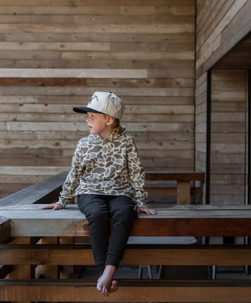 A child sitting on a wooden deck wearing a beige and brown giraffe-print hoodie, dark pants, and a beige cap with a black brim, set against a wooden wall background.
