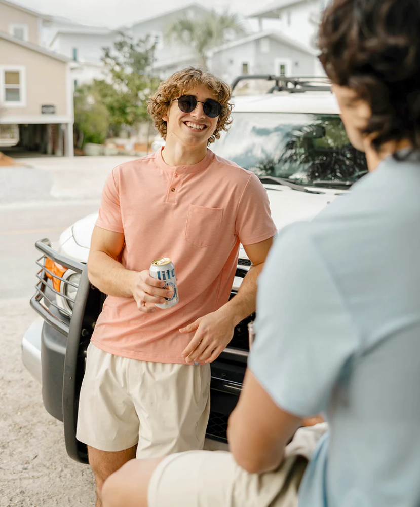 Two men standing next to a car, one holding a can wearing a Pink short-sleeve henley shirt, in a suburban setting.