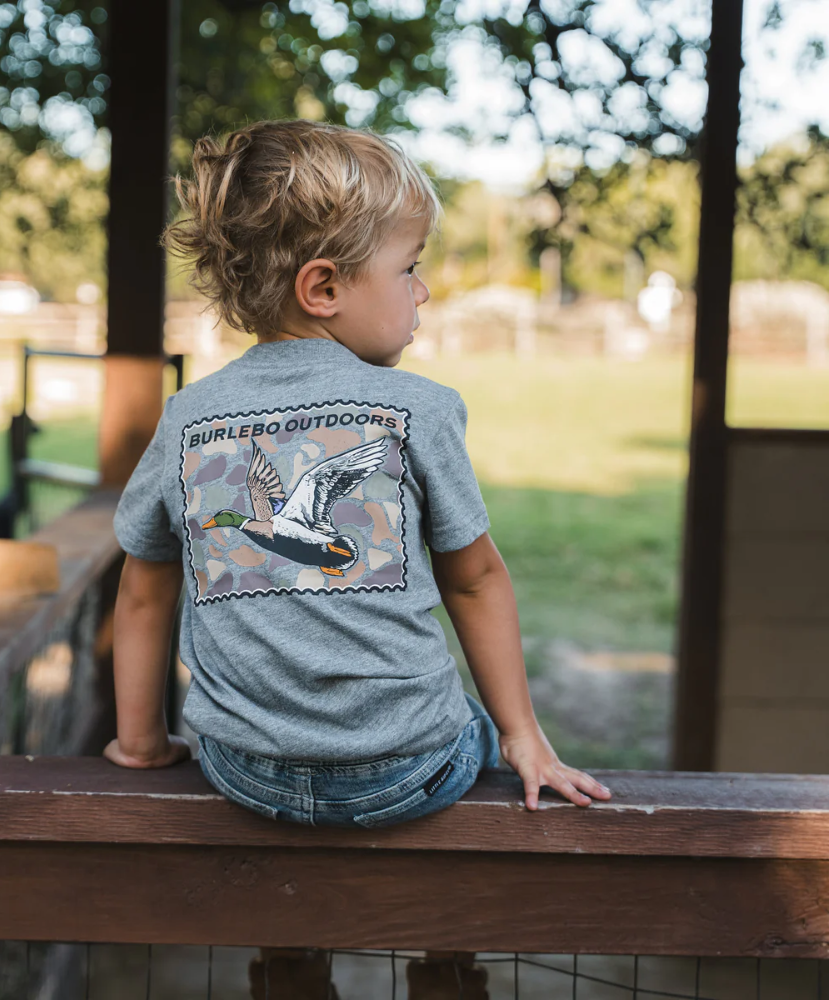Child wearing a shirt with a duck stamp graphic and text, sitting on a wooden railing outdoors.
