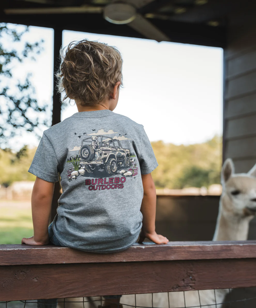 Child wearing a 'Burlebo Outdoors' t-shirt sitting on a wooden bench with an alpaca in the background.