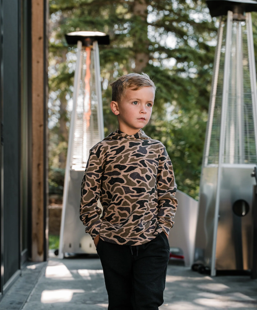 A young boy wearing a brown and black leopard print long-sleeve shirt and black pants, standing on a patio with outdoor heaters and trees in the background.