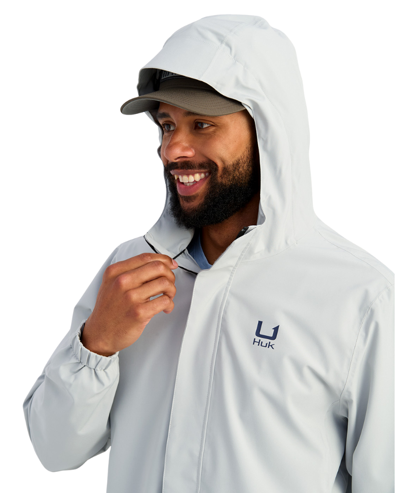 Man wearing a light gray HUK jacket and cap with a logo on a white background