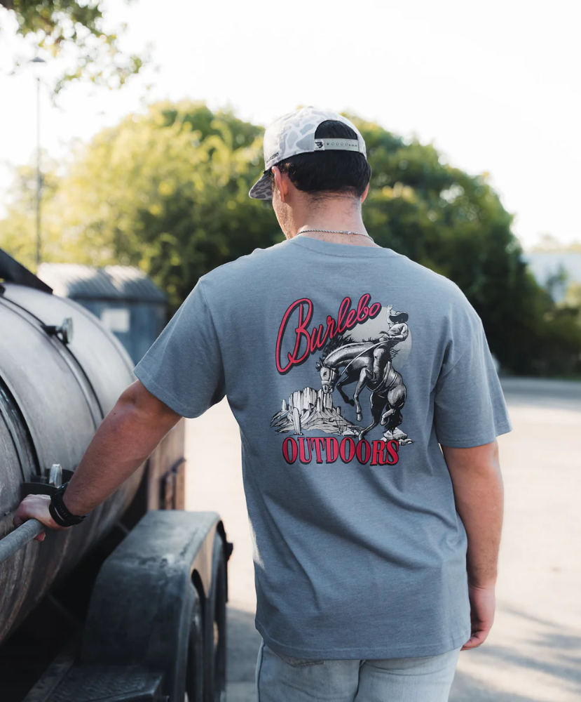 Man wearing a gray t-shirt with a graphic design and text, standing next to a barbecue grill outdoors.