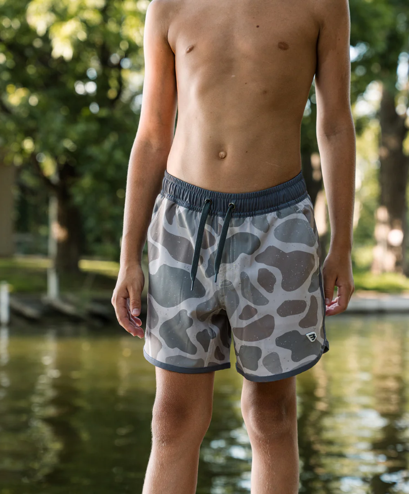 Child wearing camouflage swim shorts standing by a body of water with trees in the background