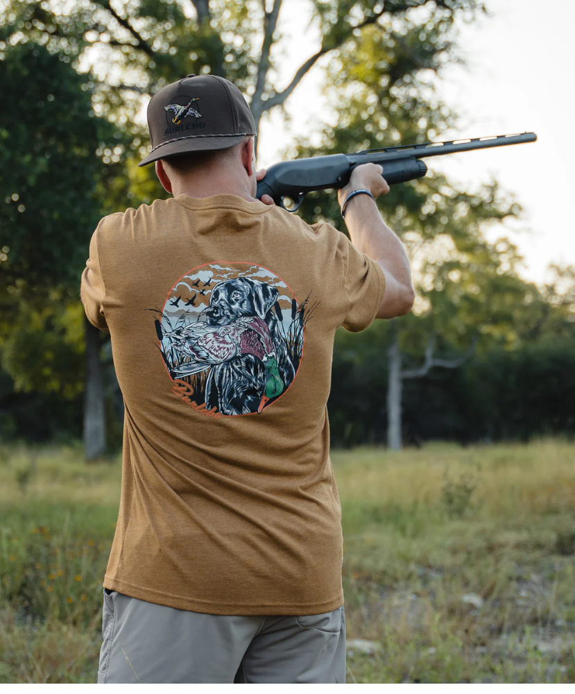Man in a brown shirt with a duck and dog design holding a shotgun outdoors.