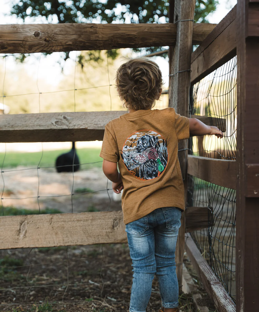 Child wearing a light brown shirt with a colorful design, standing near a wooden fence.