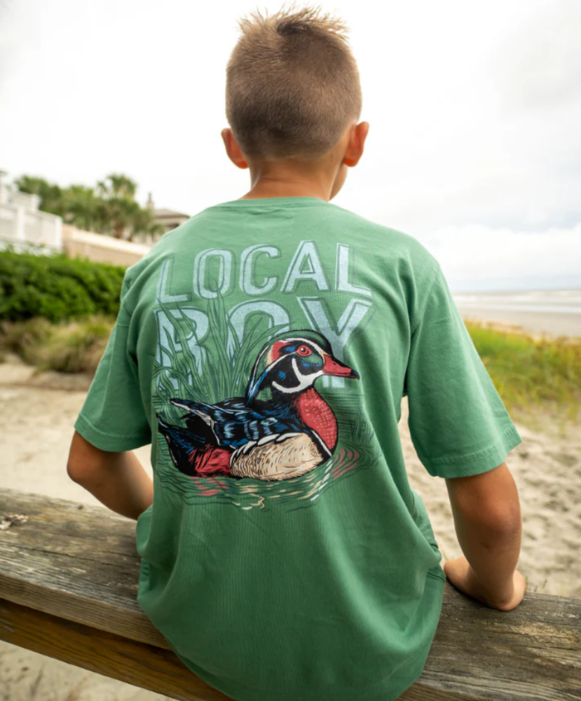 Child wearing a green t-shirt with a wood duck design and 'Local Boy' text, sitting on a wooden bench outdoors.