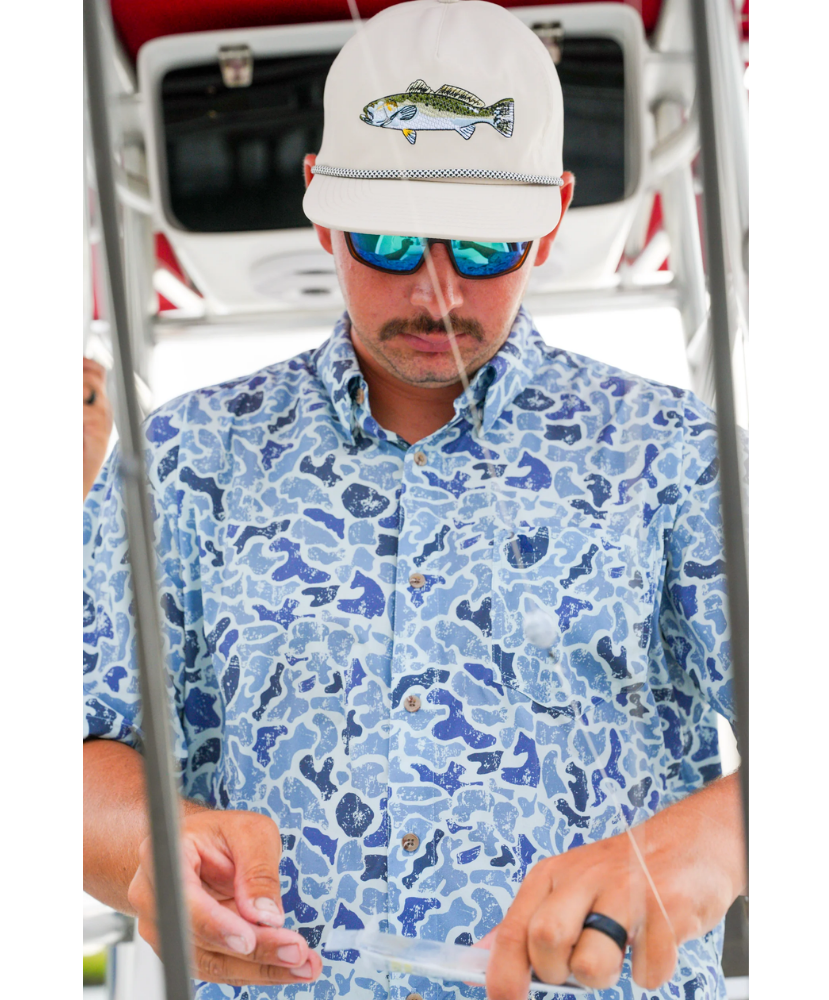Man wearing a blue patterned shirt and white cap with fish design, standing next to a boat.