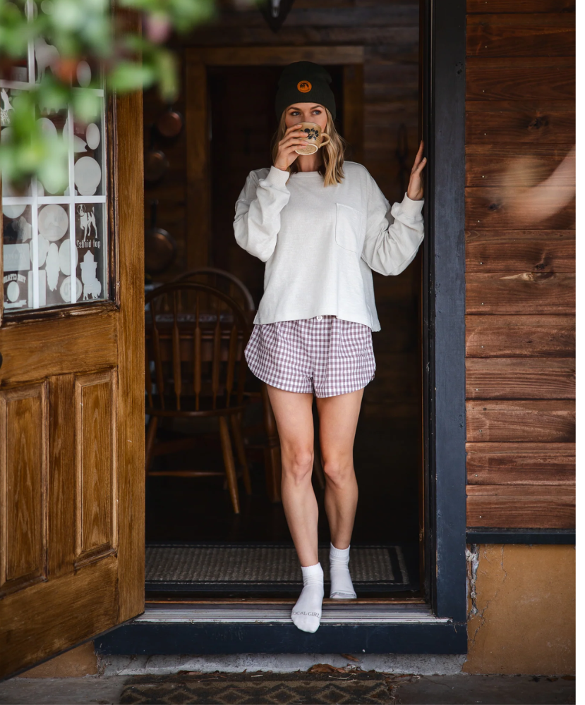 Person standing in a doorway wearing a white sweatshirt, checkered shorts, and a black cap.