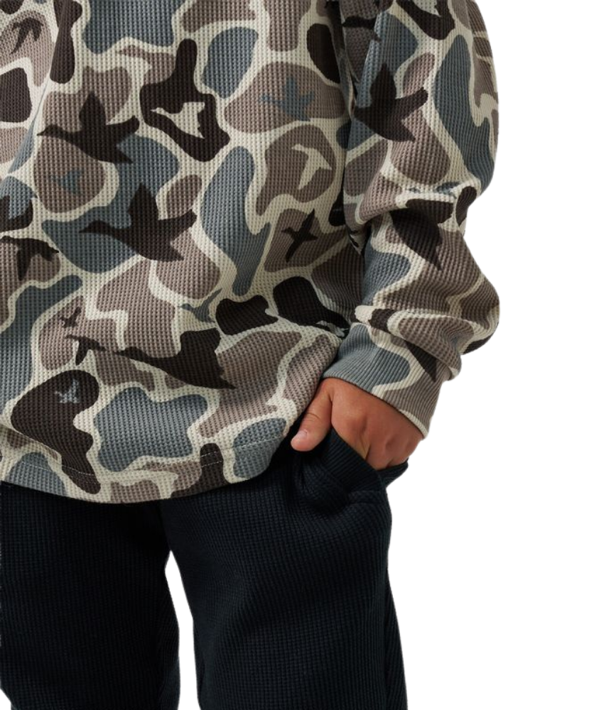 Close-up of a child wearing a waffle-knit long-sleeve sweatshirt with a muted camouflage pattern in gray, beige, brown, and black, paired with dark pants, against a white background.