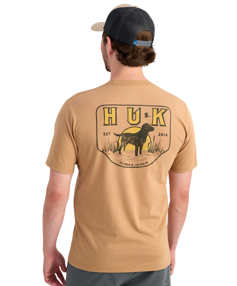 Man wearing a tan HUK shirt with a logo on the back, standing against a white background