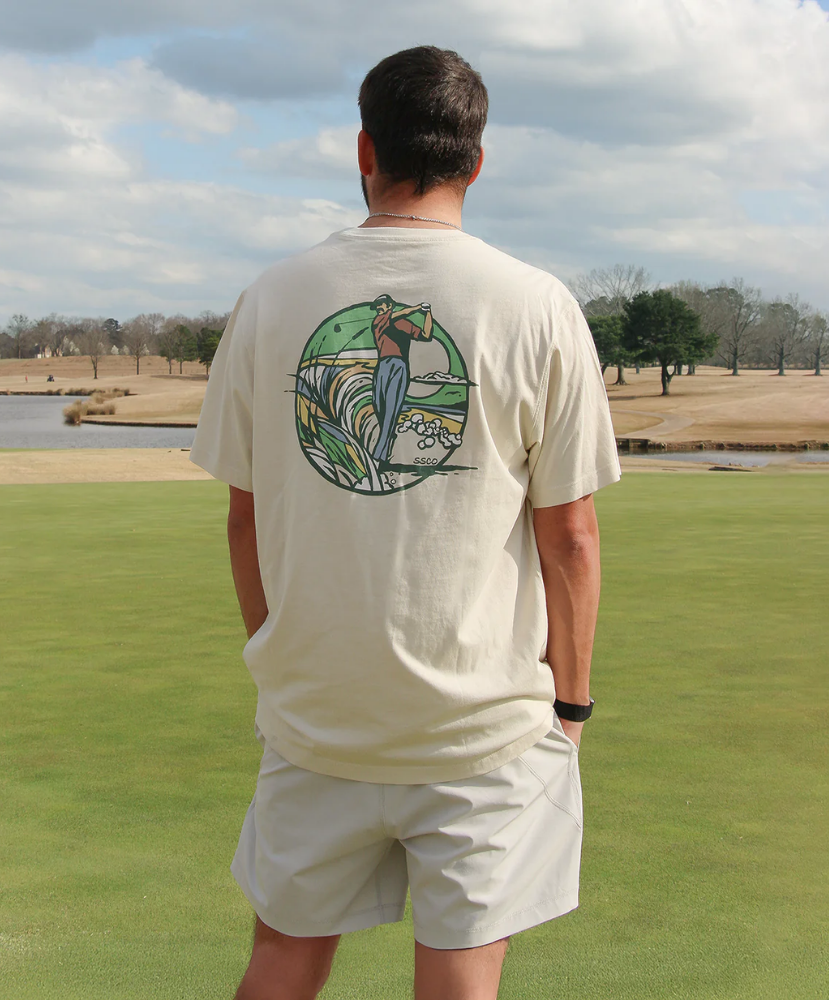 Man wearing a beige t-shirt with a GOLF graphic design on the back, standing on a golf course.