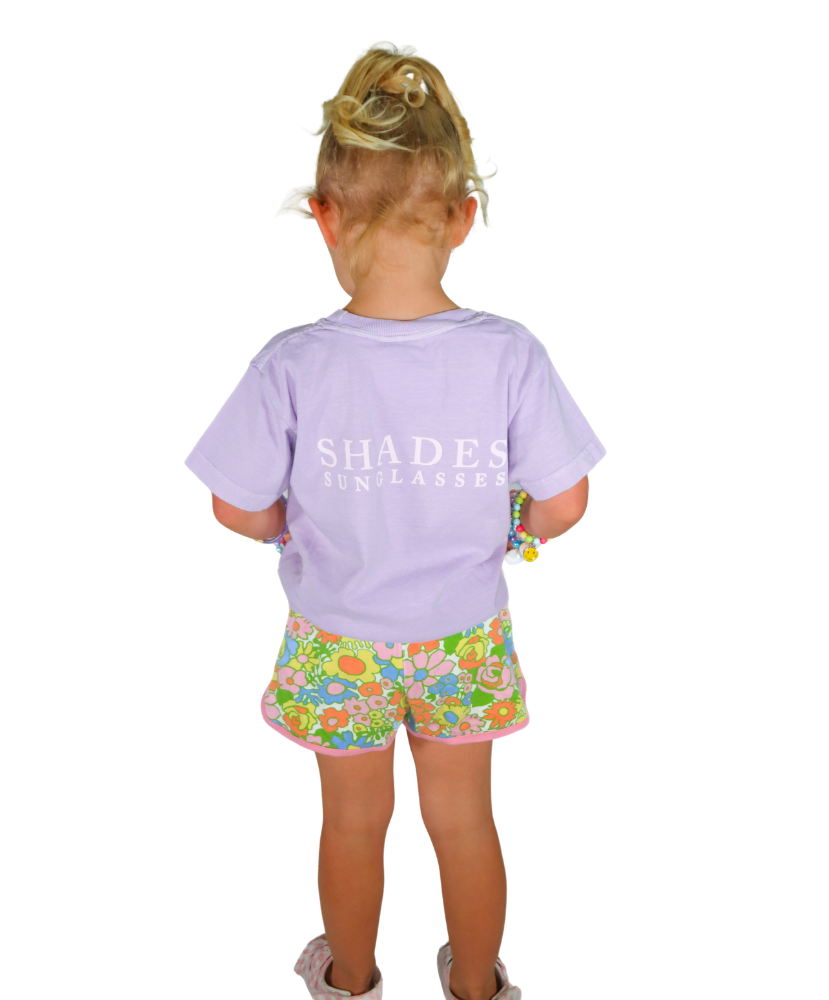 Child wearing a purple shirt with 'SHADES' text and floral shorts on a white background