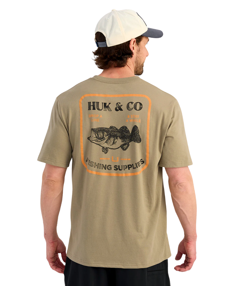 Man wearing a beige t-shirt with Huk & Co fishing supplies logo on the back. Bass graphic.