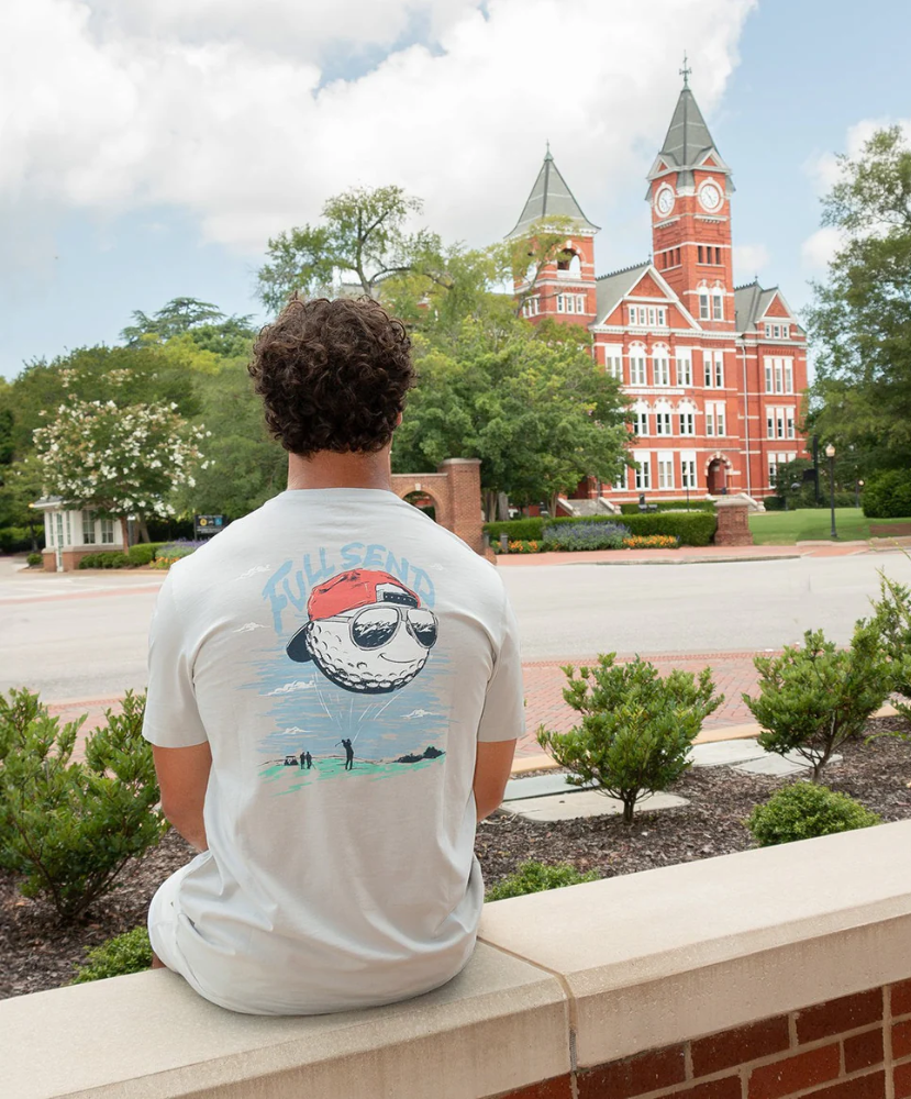 Person wearing a t-shirt with a graphic design, sitting on a ledge with a clock tower in the background.