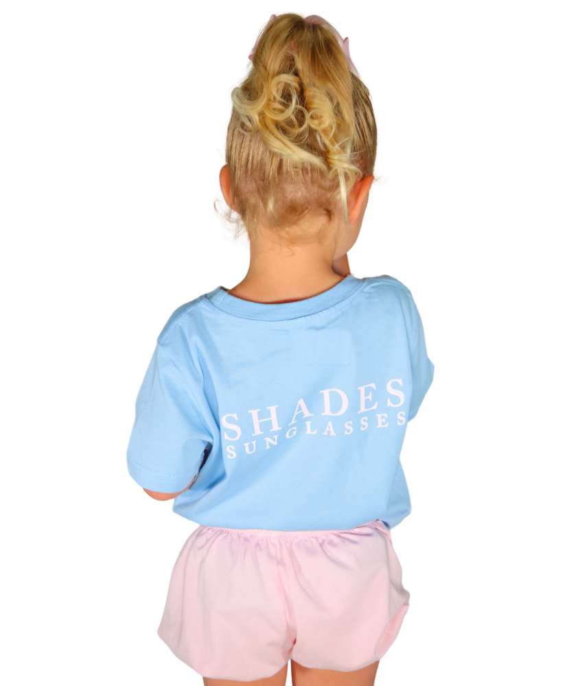 Child wearing a light blue shirt with 'SHADES SUNGLASSES' text and pink shorts on a white background