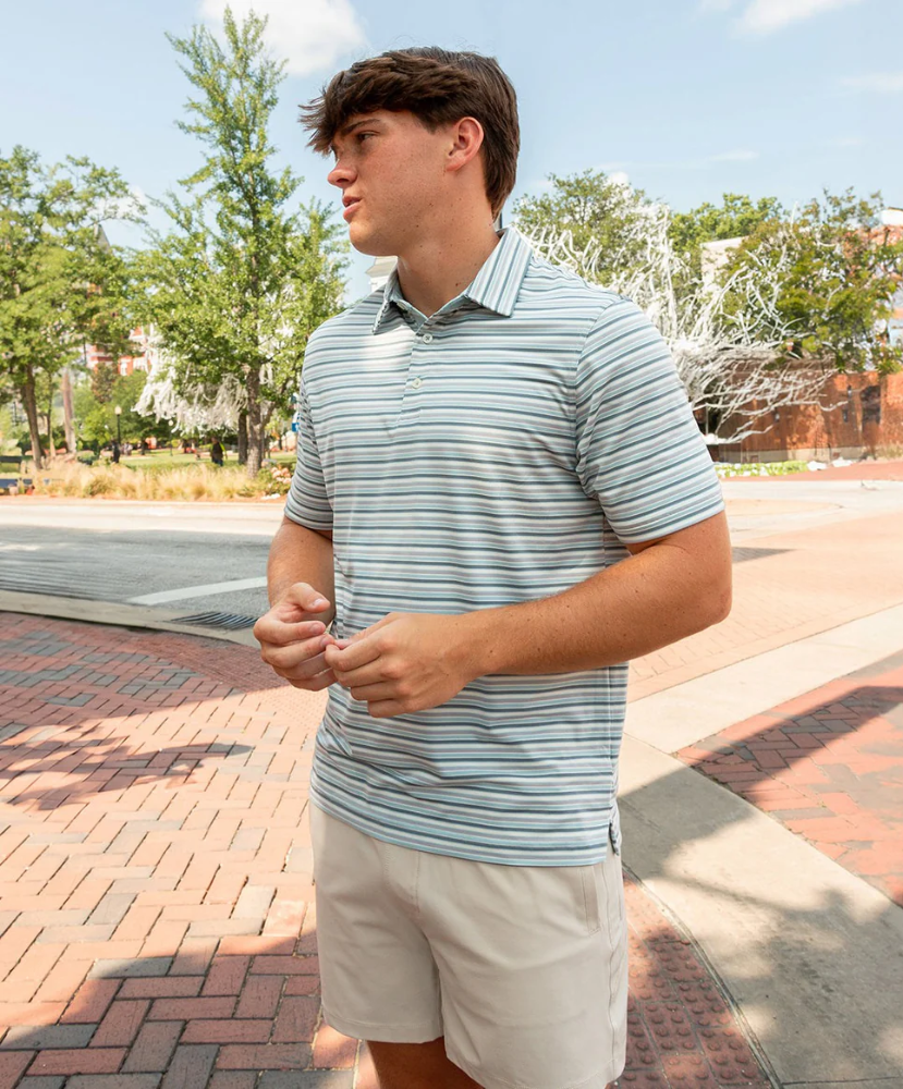 Man wearing a gray, blue, and purple striped polo shirt and shorts standing on a brick sidewalk with trees and a building in the background.
