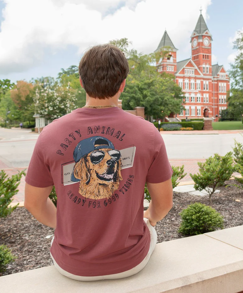 Person wearing a maroon t-shirt with a dog graphic and text, sitting in front of a red brick building.