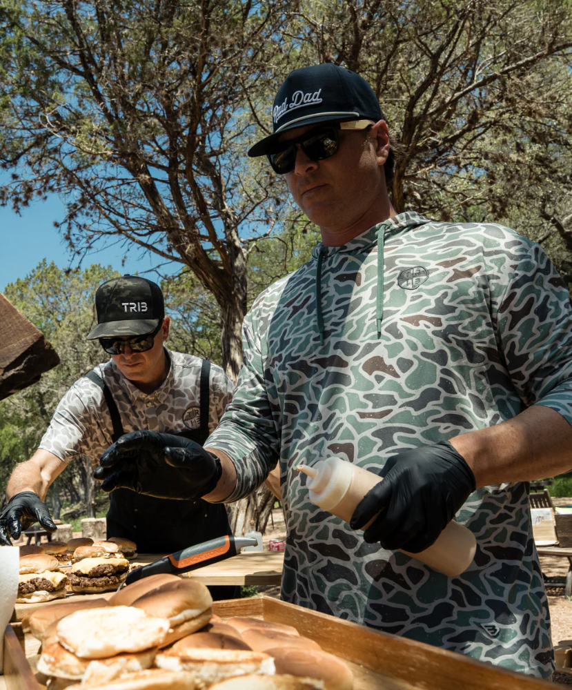 Two men preparing food outdoors, with one wearing a retro duck camoflage hoodie