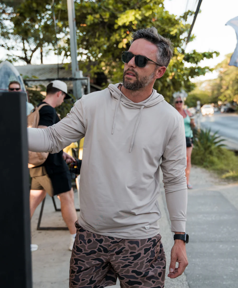 Man wearing a BEIGE hoodie and camouflage shorts outdoors with trees and people in the background.
