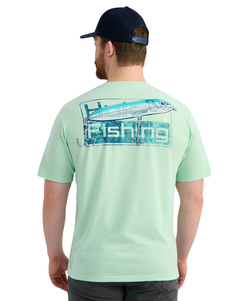 Man wearing a light green HUK t-shirt with a fish lure design and 'Fishing' text, standing against a white background.