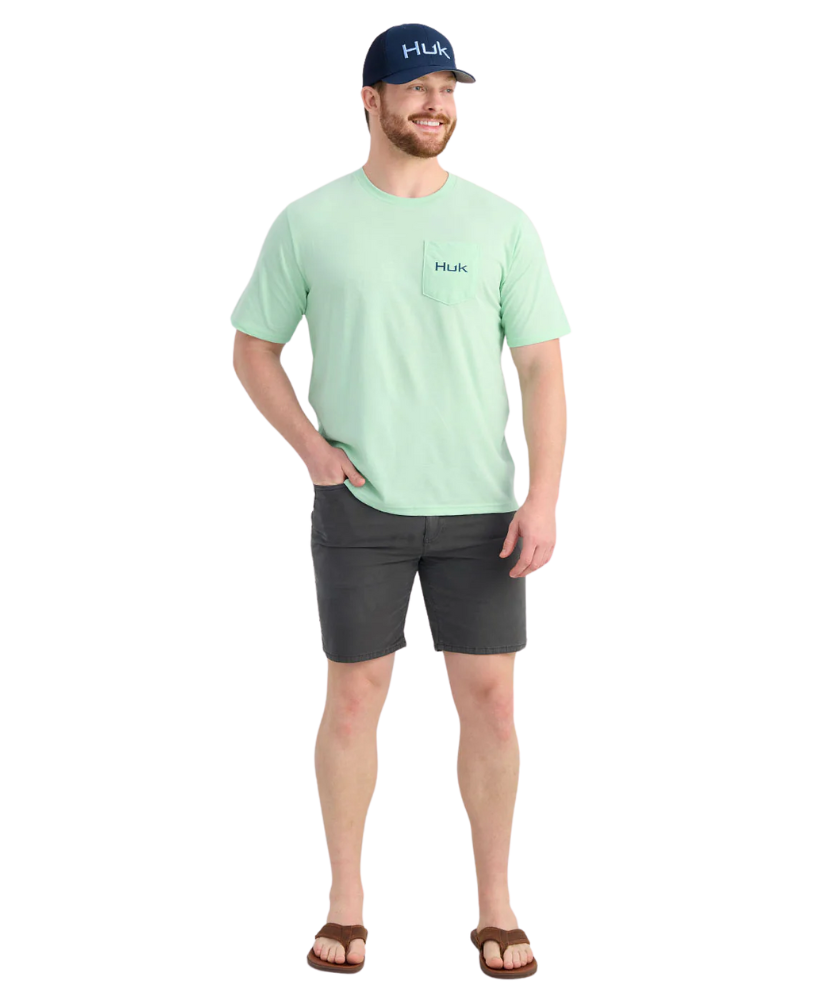 Man wearing a light green HUK t-shirt and dark shorts with a white background