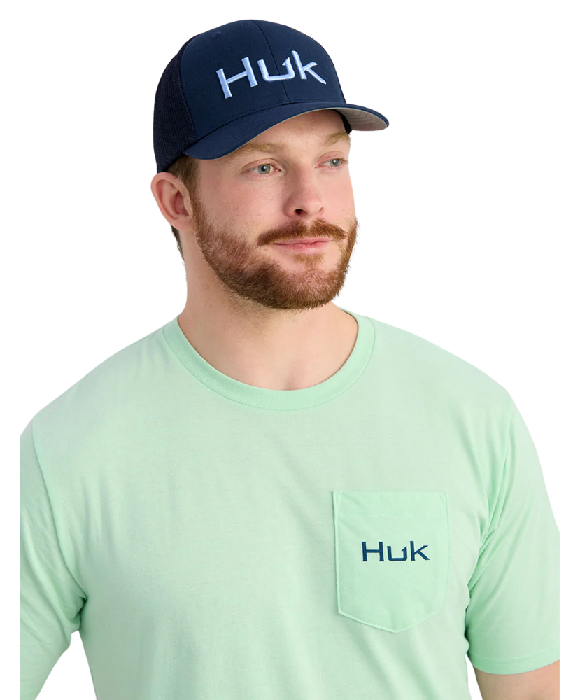 Man wearing a green Huk shirt and navy cap on a white background