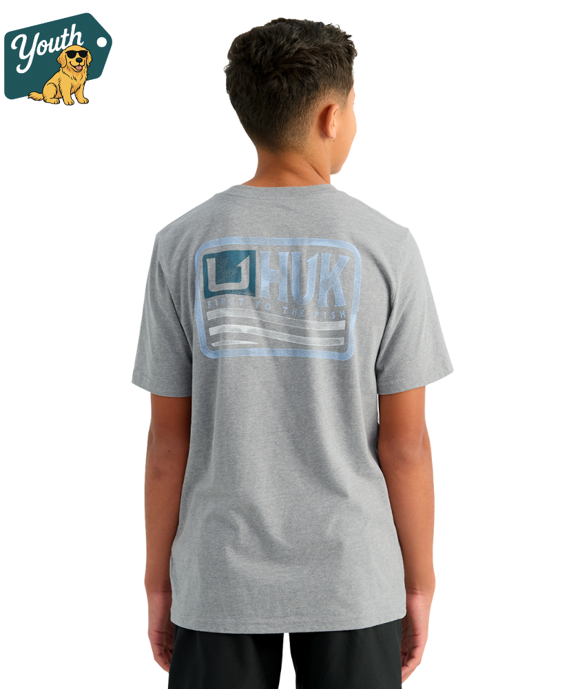 KID wearing a gray HUK t-shirt with a logo on the back, standing against a white background.