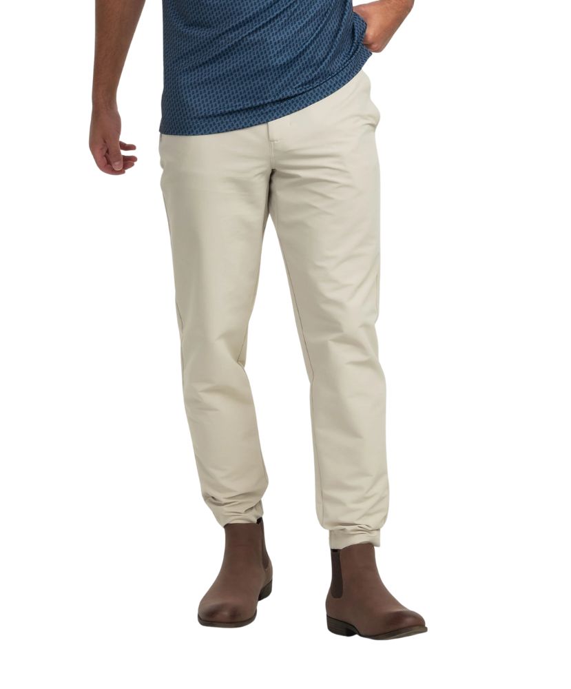 A man wearing beige Nomad performance joggers with a pair of boots and a blue shirt. The joggers have an elastic waistband with belt loops and feature side and back pockets.