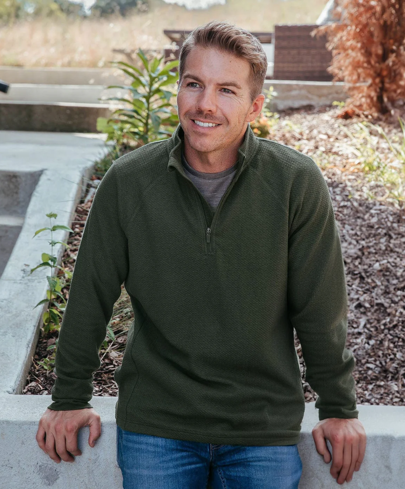Man wearing a green pullover standing outdoors with plants and a building in the background