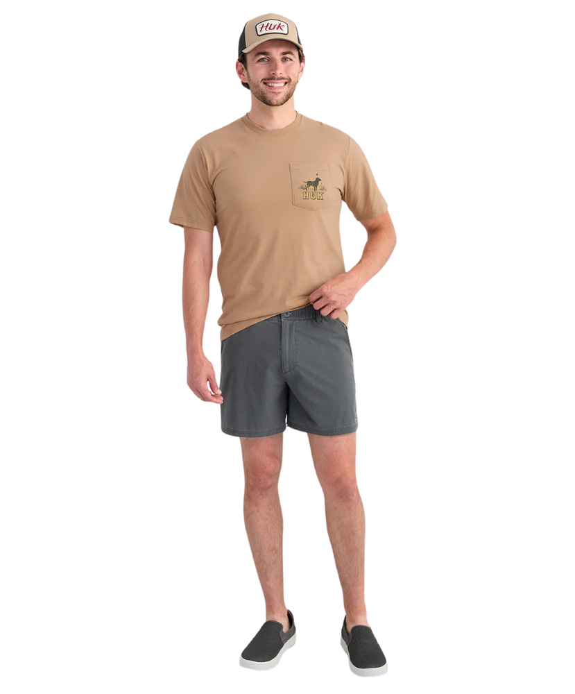 Man wearing a tan t-shirt with a logo and gray HUK shorts on a white background