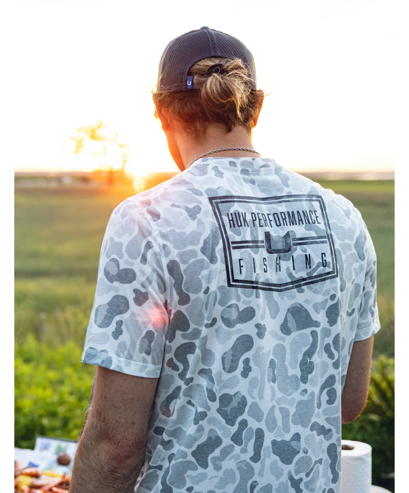 Man wearing a camouflage t-shirt with 'Nuk Performance Fishing' logo, standing outdoors with sunset in the background.