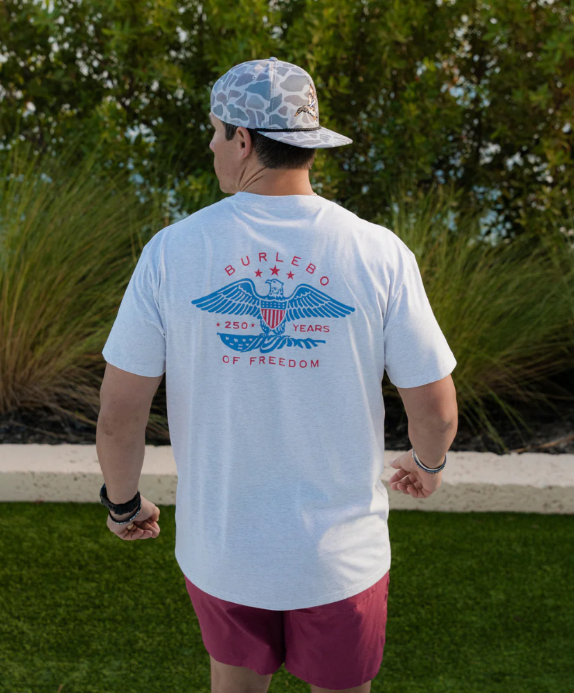 Person wearing a white t-shirt with a graphic design, standing outdoors with greenery in the background.