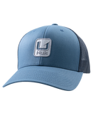 A blue trucker-style baseball cap with a structured front panel, curved brim, and dark mesh back panels. A square embroidered patch on the front displays a stylized 'H' logo above the word 'Huk', indicating the brand.