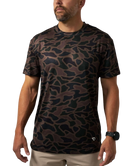 A man wearing a short-sleeve crewneck t-shirt with a dark brown and black organic camouflage pattern, paired with khaki pants and a black watch, against a plain white background.