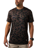A man wearing a short-sleeve crewneck t-shirt with a dark brown and black organic camouflage pattern, paired with khaki pants and a black watch, against a plain white background.