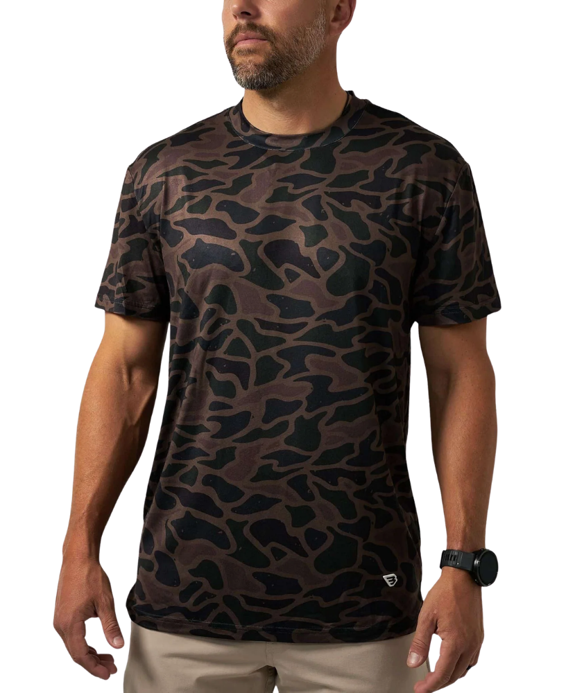 A man wearing a short-sleeve crewneck t-shirt with a dark brown and black organic camouflage pattern, paired with khaki pants and a black watch, against a plain white background.