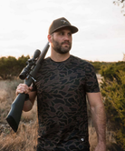 A man wearing a dark brown and black camouflage t-shirt and a matching baseball cap, holding a rifle with a scope over his shoulder in an outdoor field setting.